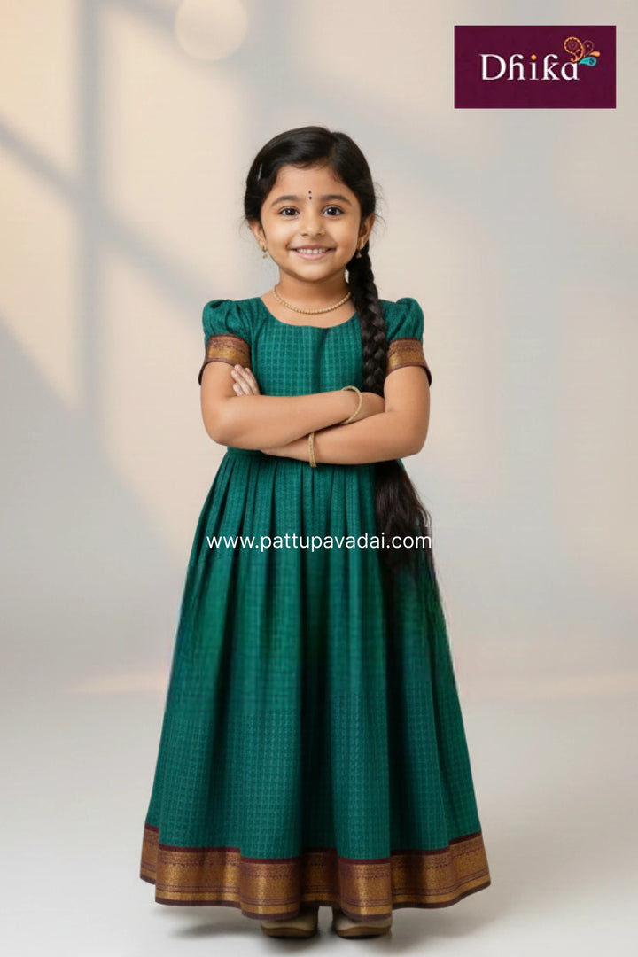 Young girl wearing a green traditional dress with brown accents, standing against a plain background with 'Dhika' branding.