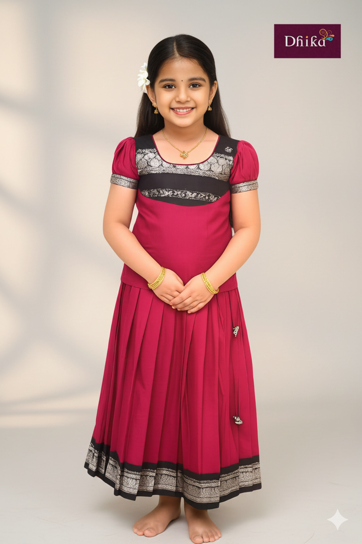 Young girl wearing a red traditional outfit with black and silver details on a plain background, branded 'Dhika'.