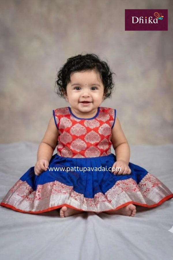 Baby wearing a red and blue dress with a textured pattern, sitting on a gray surface.