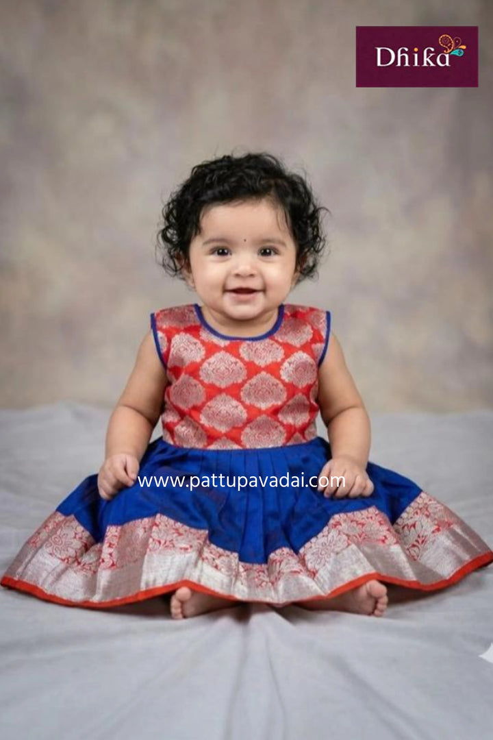 Baby wearing a red and blue dress with a textured pattern, sitting on a gray surface.