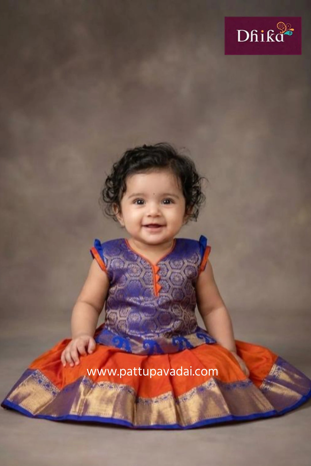 Child wearing a colorful traditional outfit with a brown background, featuring the brand 'Dhika'.
