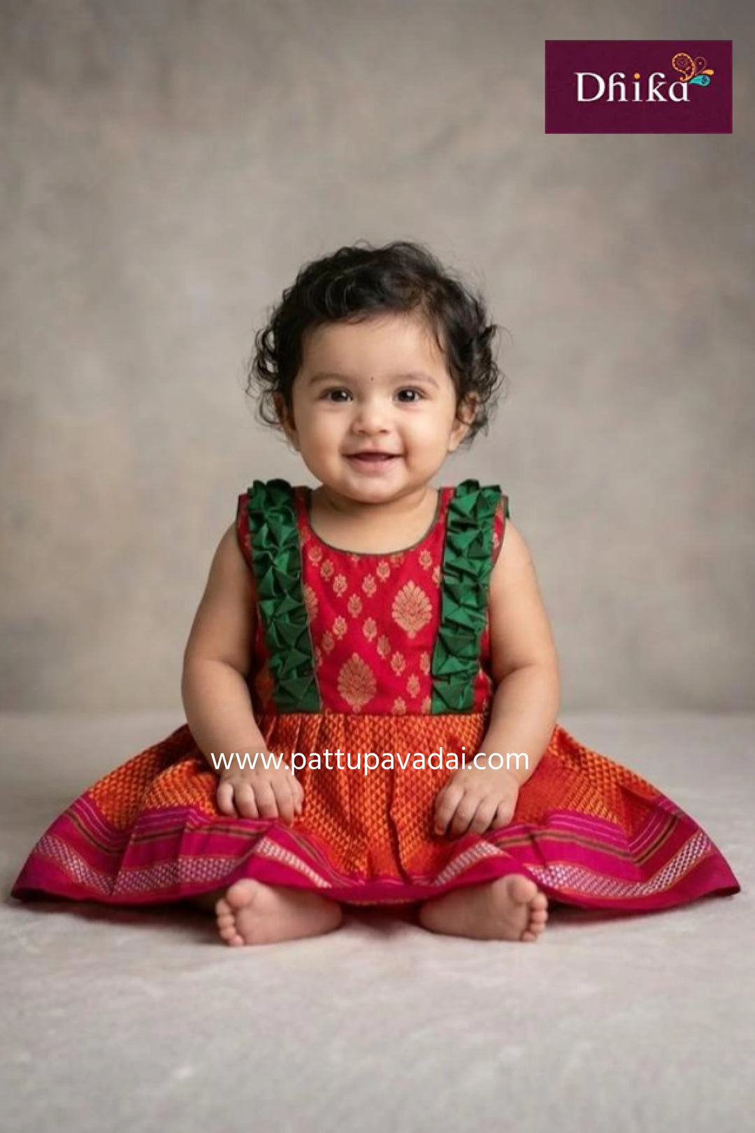 Child wearing a red and orange traditional outfit with green ruffles, sitting on a plain background.