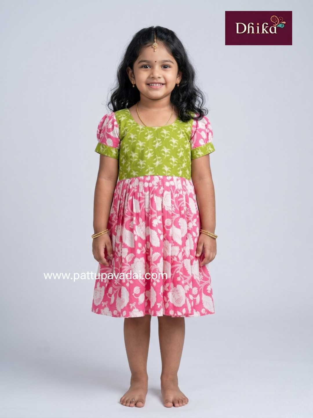 Young girl wearing a green and pink dress with floral patterns on a white background, featuring the brand 'Dhika'.