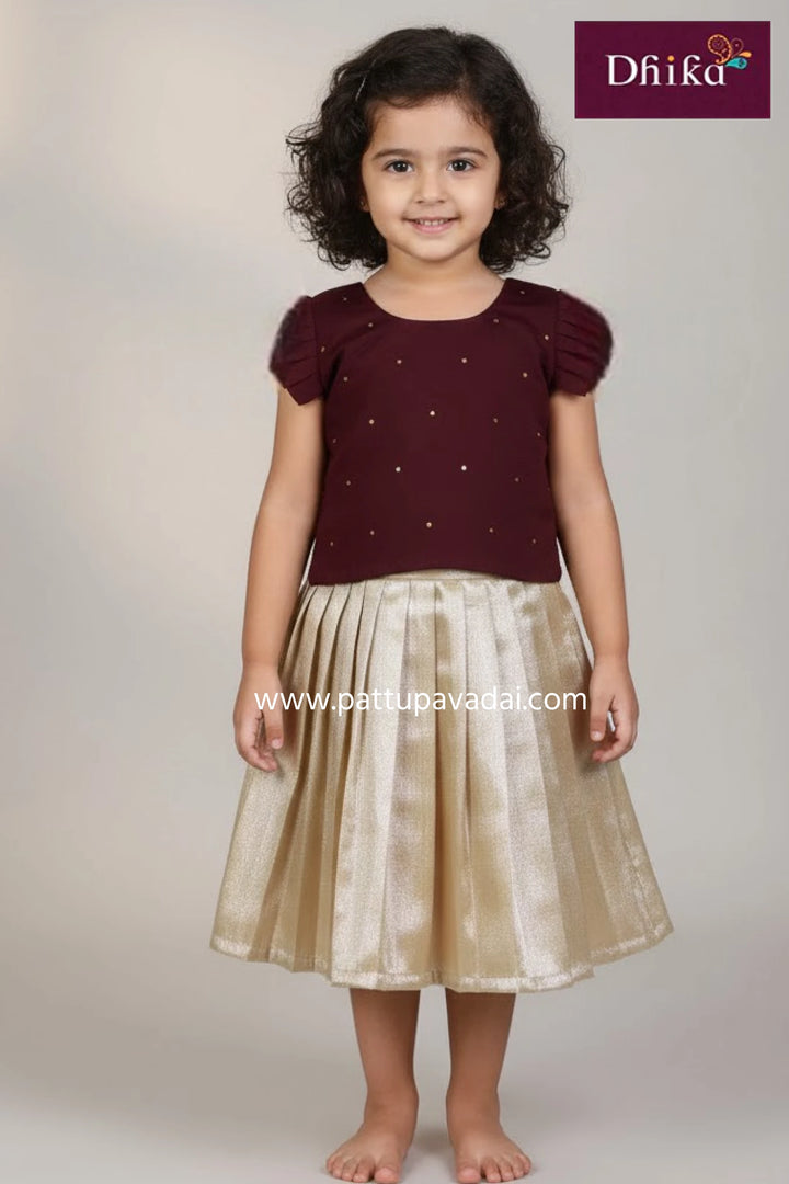 Young girl wearing a maroon top with short sleeves and a gold skirt, standing against a plain background. The brand 'Dhika' is visible.
