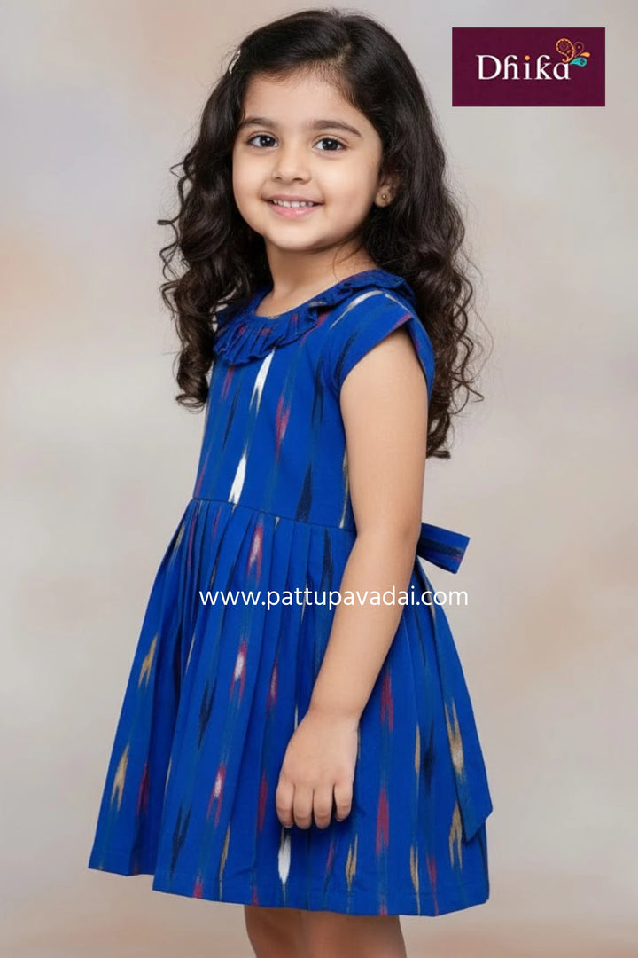 Young girl wearing a blue dress with a pattern, standing against a plain background.
