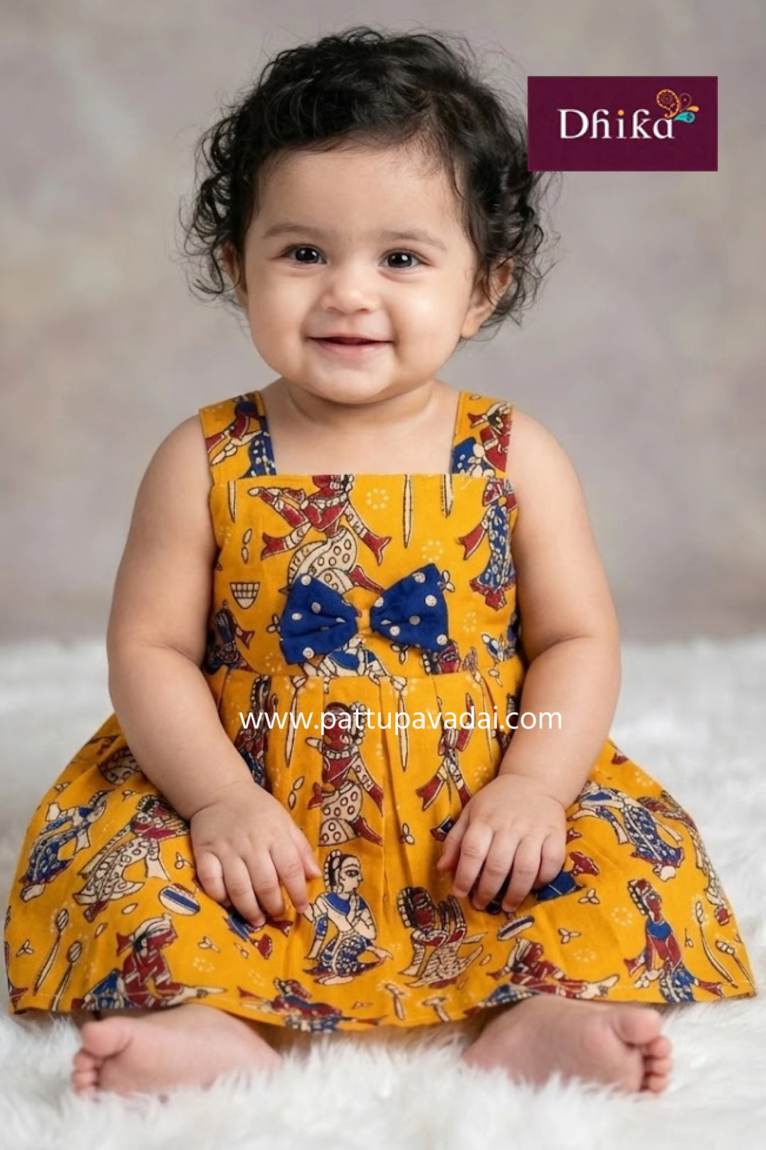 Child wearing a yellow dress with colorful patterns and a blue bow, sitting on a white surface with a gray background.
