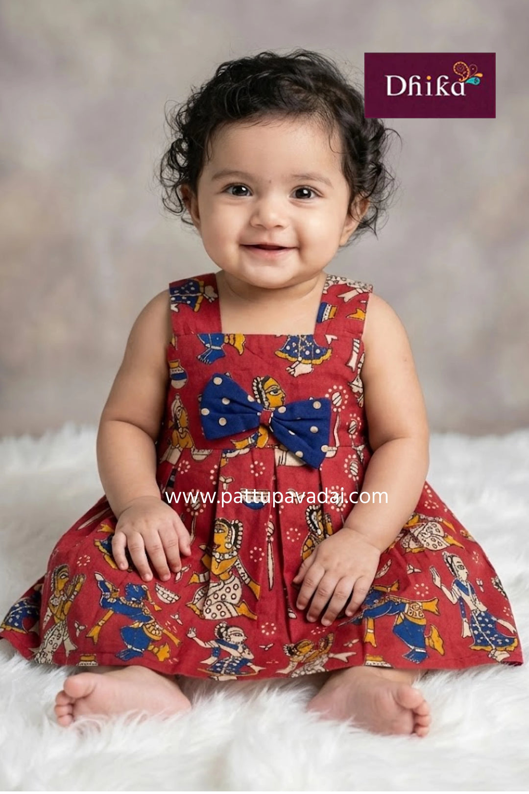 Baby wearing a red dress with colorful patterns and a blue bow, sitting on a white surface with a gray background.