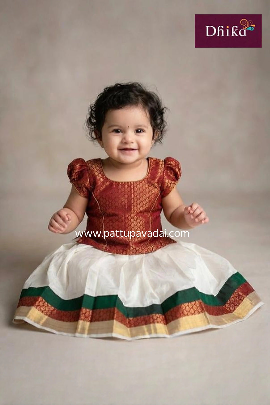 Baby wearing a traditional outfit with a plain background, featuring the brand 'Dhika'.
