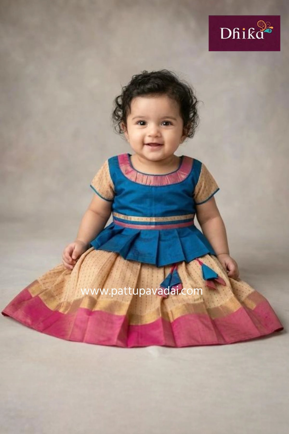 Child wearing a traditional outfit with blue and pink top and beige skirt, brand 'Dhika' visible.