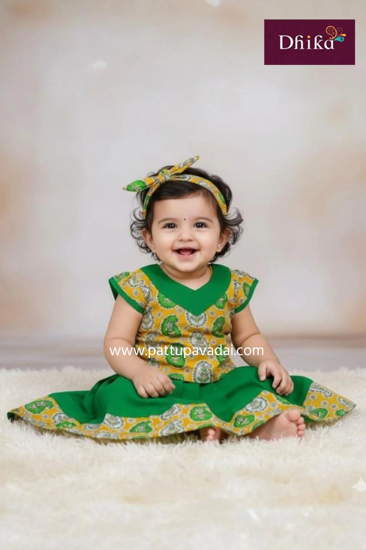 Baby wearing a green and yellow dress with a matching headband, sitting on a white surface.