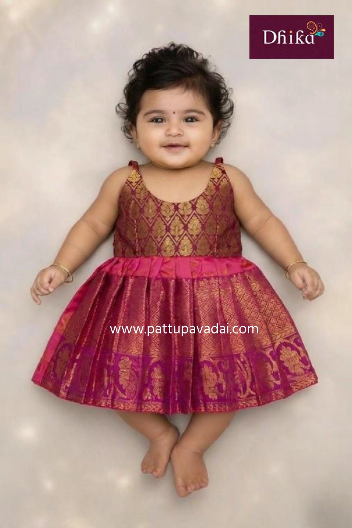 Baby wearing a pink and gold traditional dress with 'Dhika' branding.