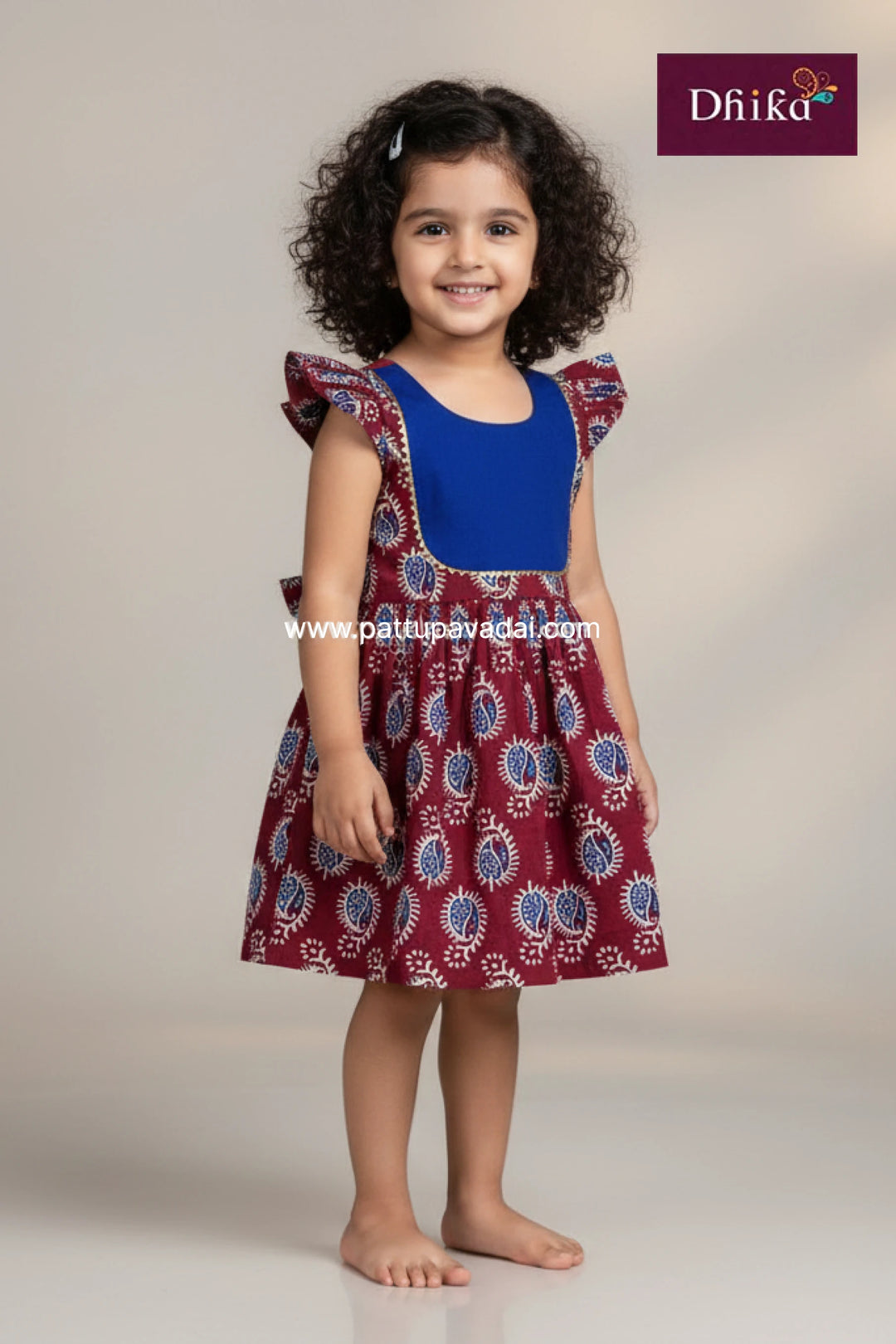 Young girl wearing a blue top and maroon patterned dress with 'Dhika' branding.