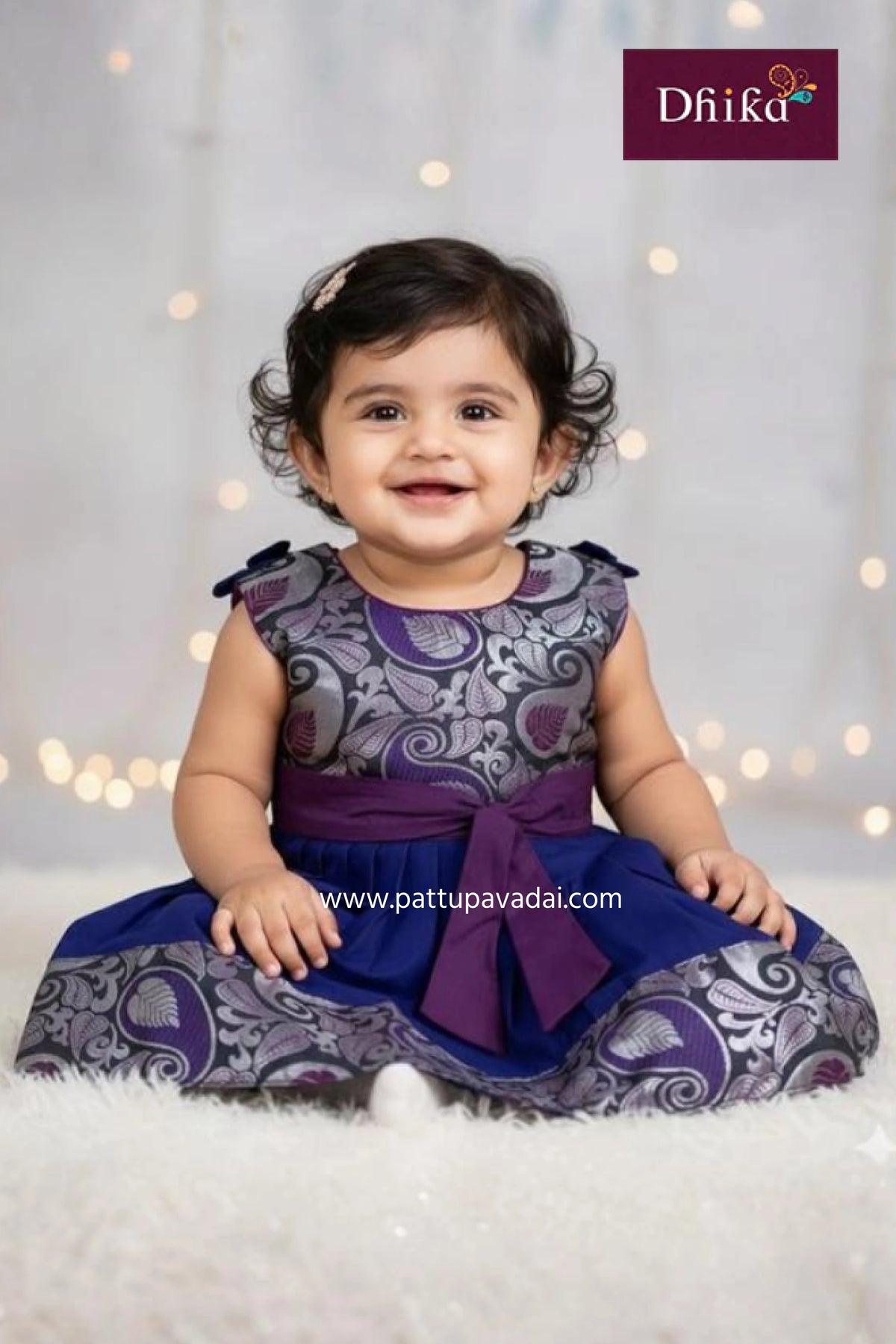 Baby wearing a patterned outfit with a purple bow, sitting on a white surface with a soft focus background.