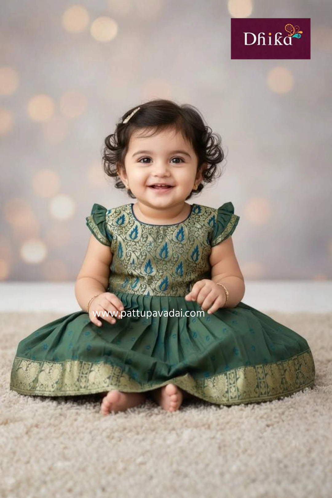 Baby wearing a green traditional outfit with a blurred bokeh background, featuring the Dhika brand.