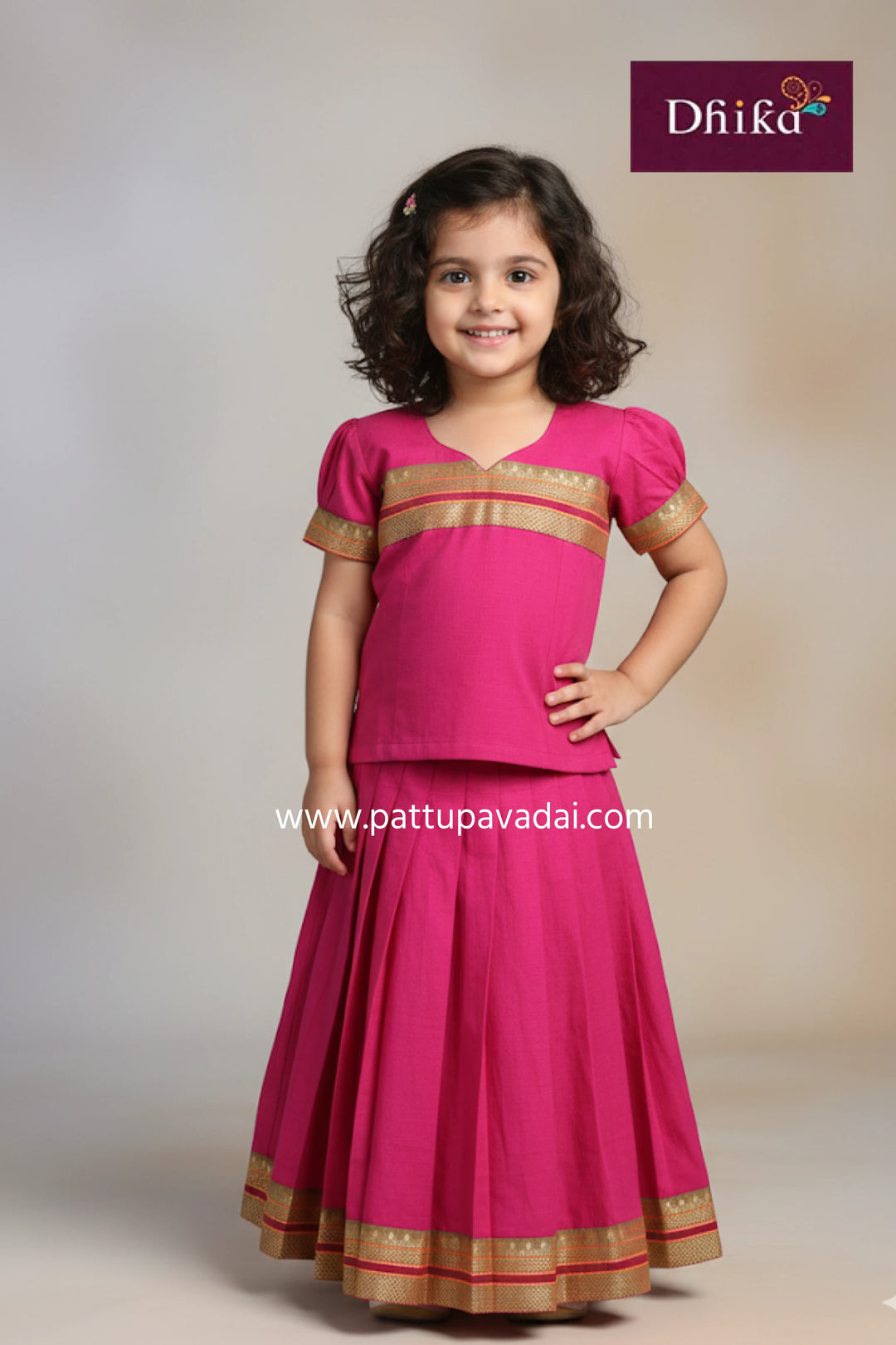 Young girl wearing a pink traditional outfit with gold accents, standing against a plain background with 'Dhika' branding.