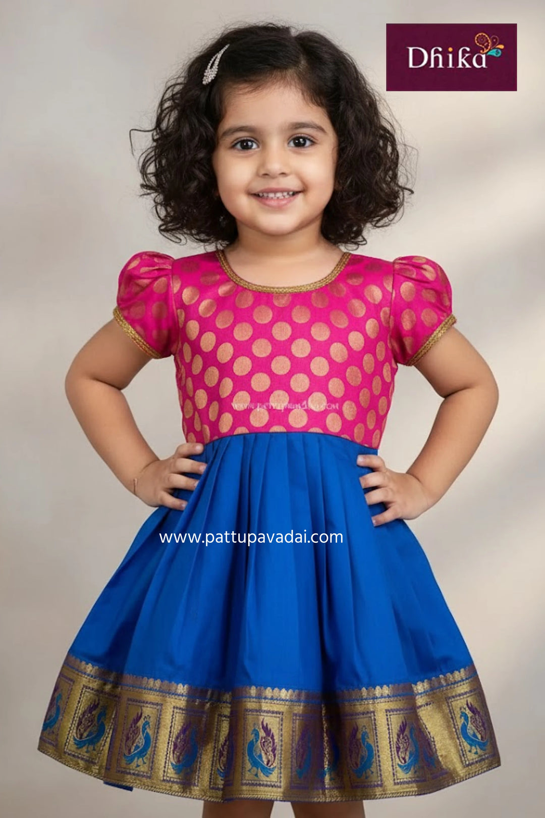 Young girl wearing a pink and blue traditional outfit with gold accents, branded 'Dhika'.