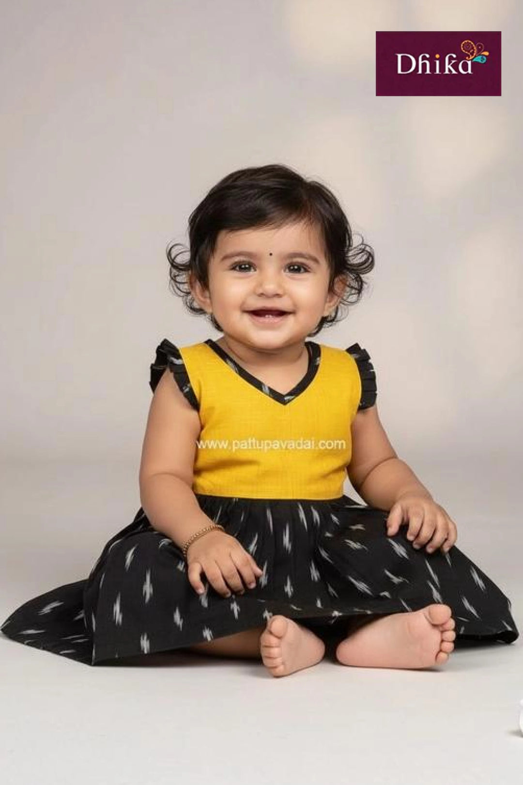 Baby wearing a yellow top and black skirt with white patterns, sitting on a white background.