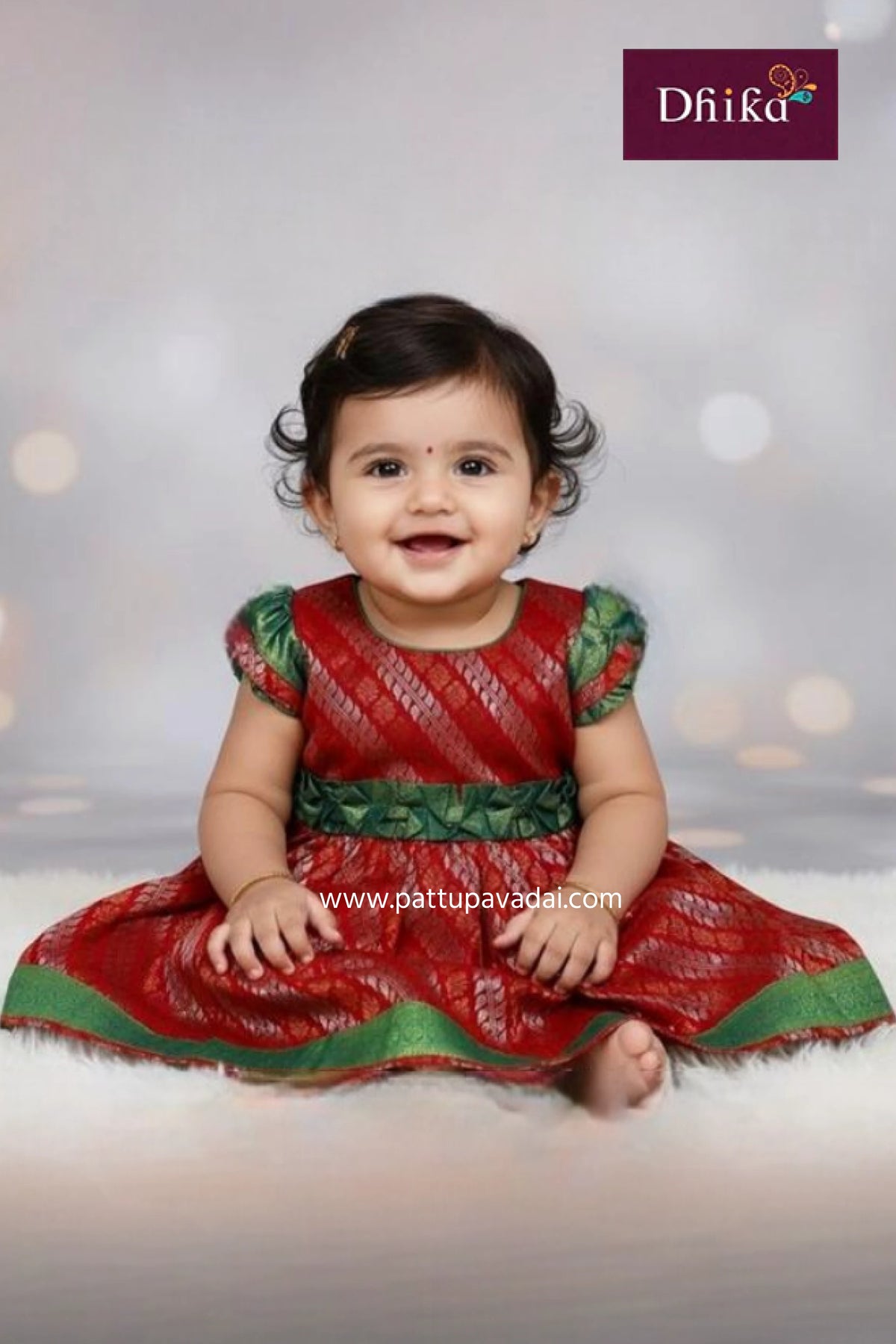 Baby wearing a red and green traditional outfit with 'Dhika' branding.