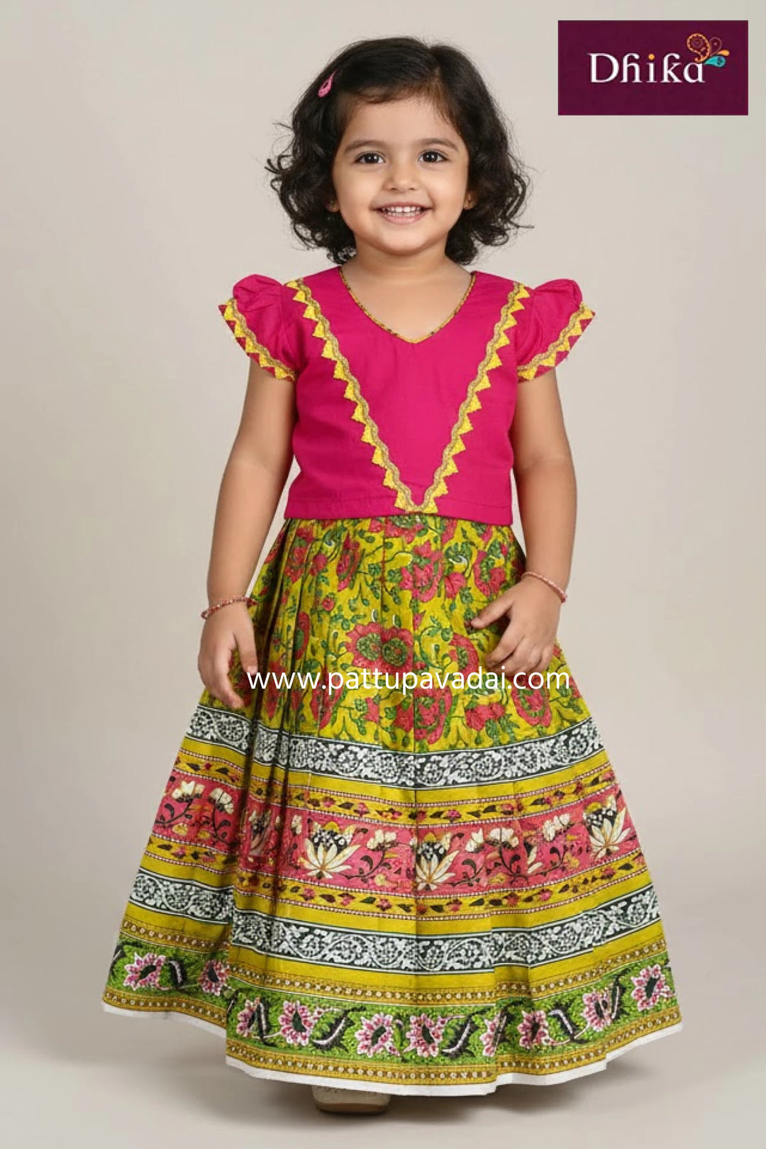 Young girl wearing a colorful traditional outfit with a pink top and patterned skirt, branded 'Dhika'.