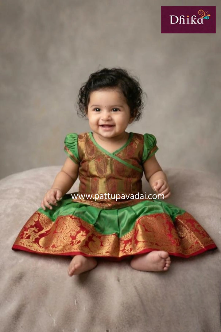 Baby wearing a traditional outfit with green and red colors on a gray background, featuring the brand 'Dhika'.