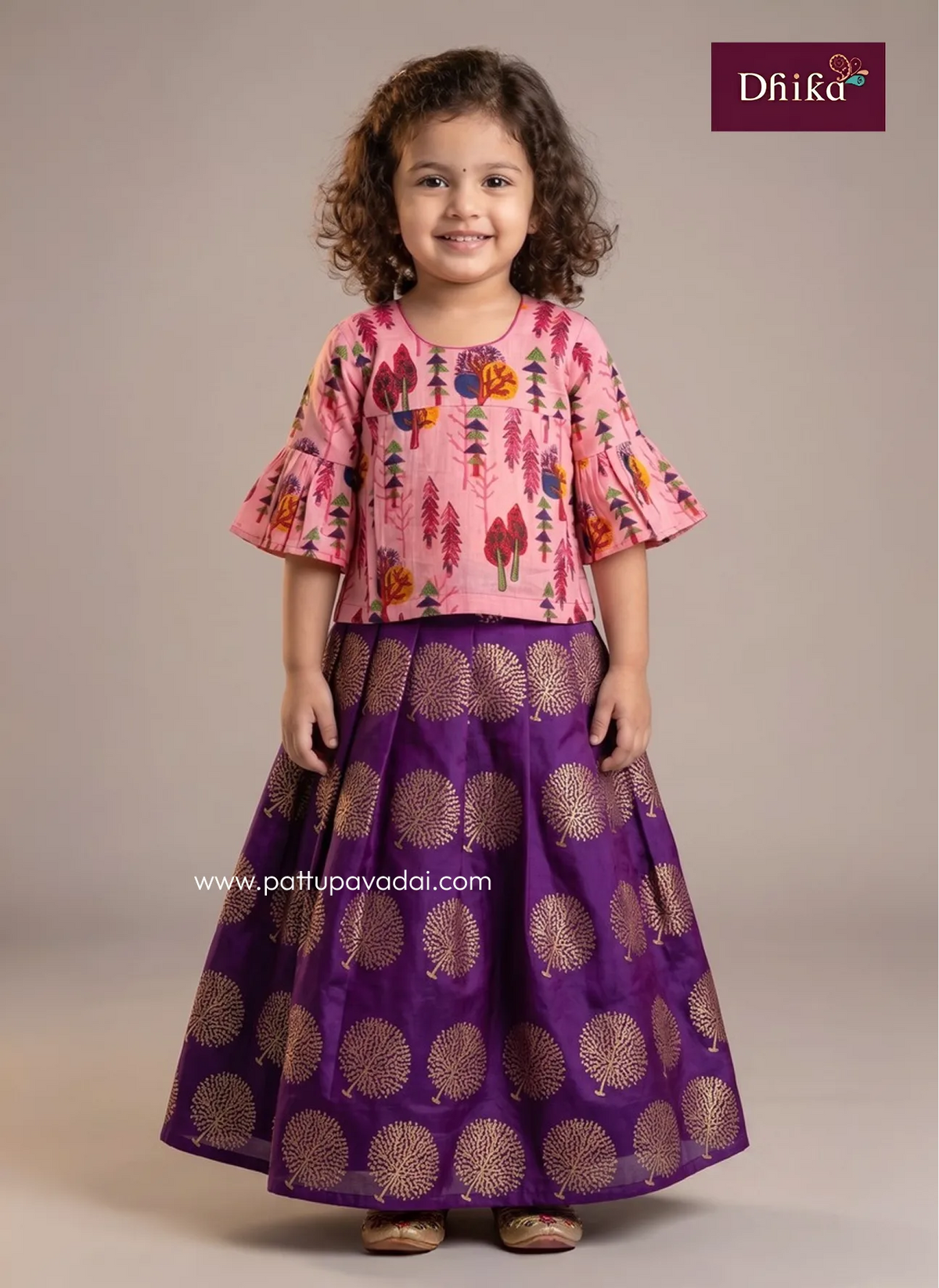 A young girl with curly brown hair stands against a plain beige studio background wearing a two-piece traditional outfit. The top is light pink with a colorful tree and floral print, and the skirt is deep purple with metallic gold circular motifs.
