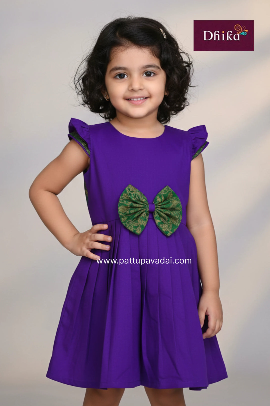 Young girl wearing a purple dress with a green bow, standing against a plain background.