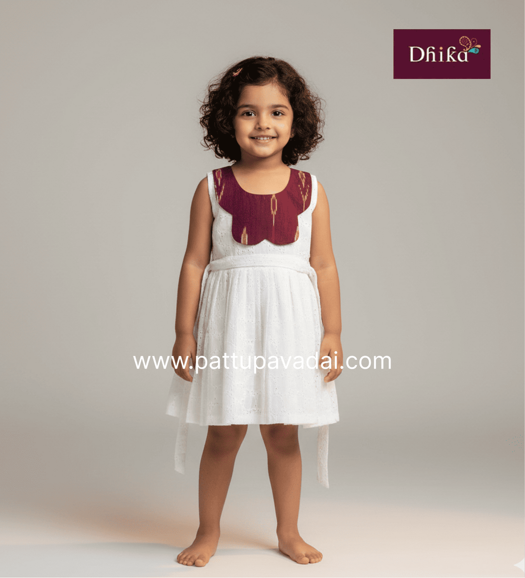 Young girl wearing a white dress with red embroidery, standing against a plain background. Dhika brand logo is visible.