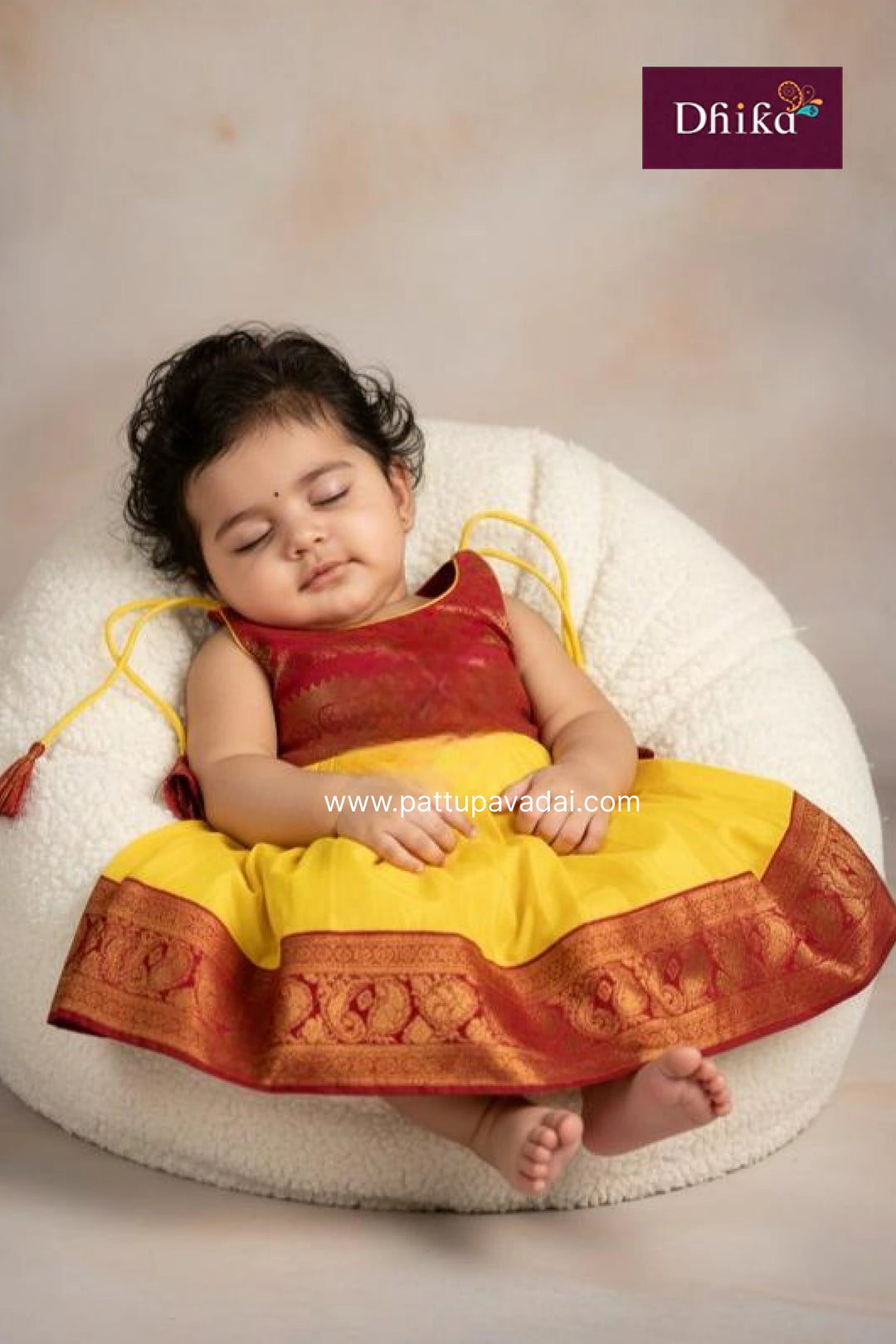Baby in traditional outfit sitting on a white cushion with 'Dhika' brand logo.