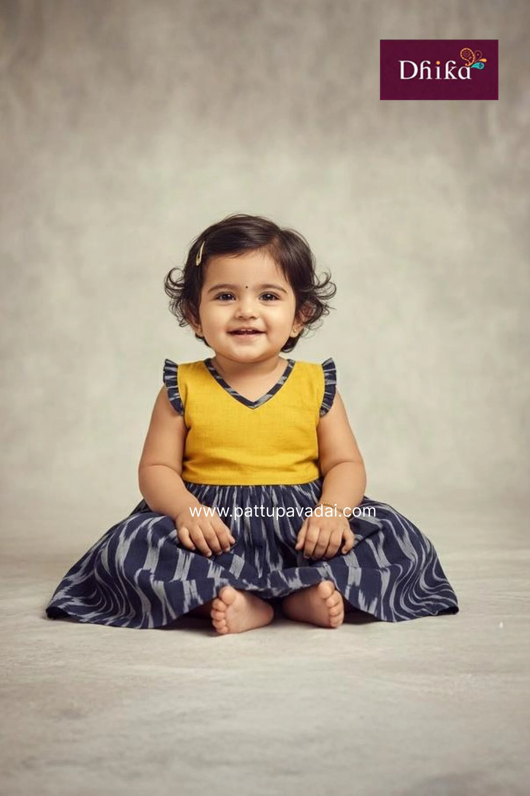 Child wearing a yellow top and blue patterned skirt sitting on a gray background with Dhika branding.