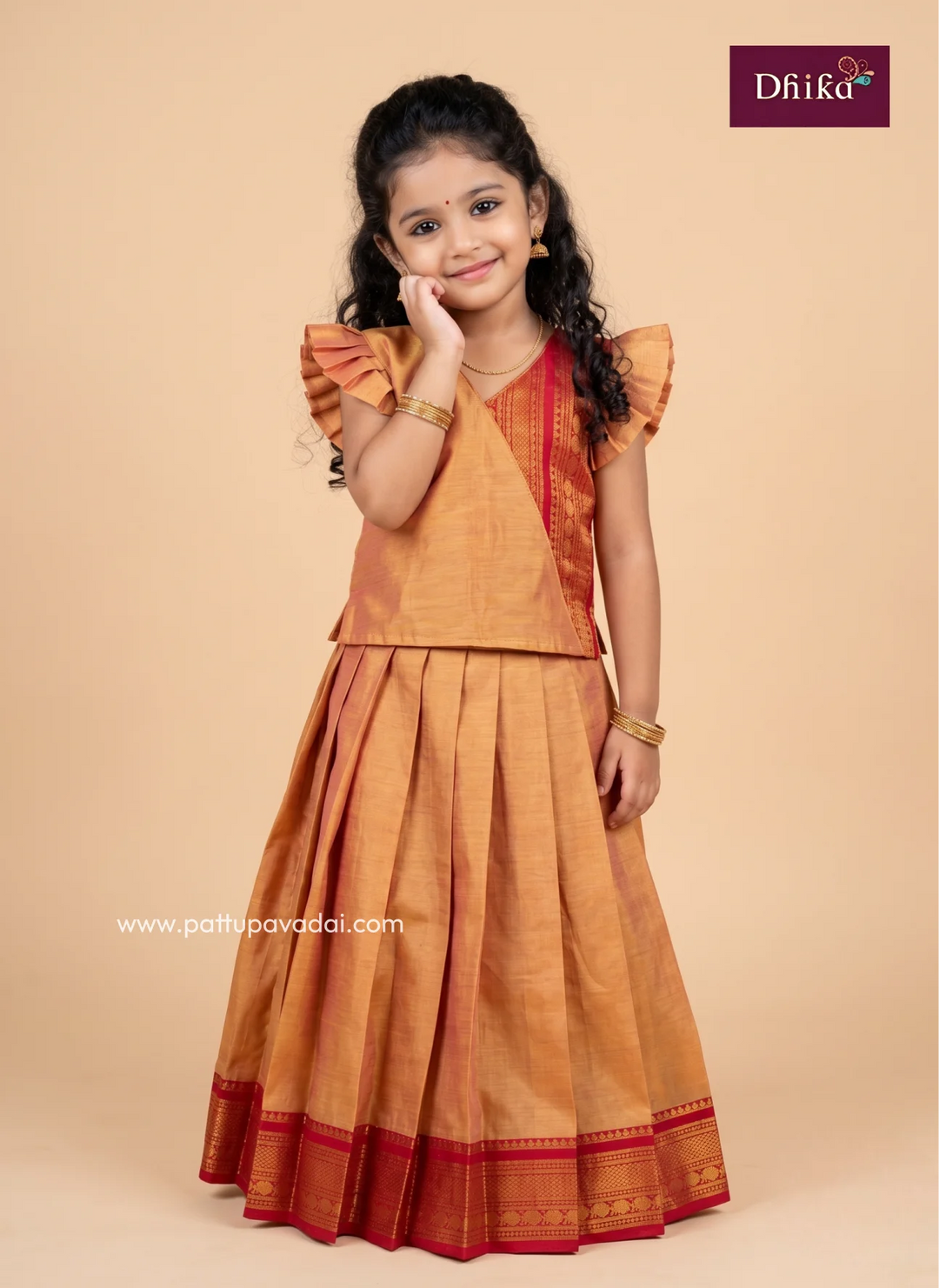 A young girl modeling a traditional Indian outfit consisting of a mustard-gold pleated skirt with red zari borders and a matching gold top with ruffled sleeves, set against a solid beige background. The brand logo 'Dhika' is visible in the top right corner.
