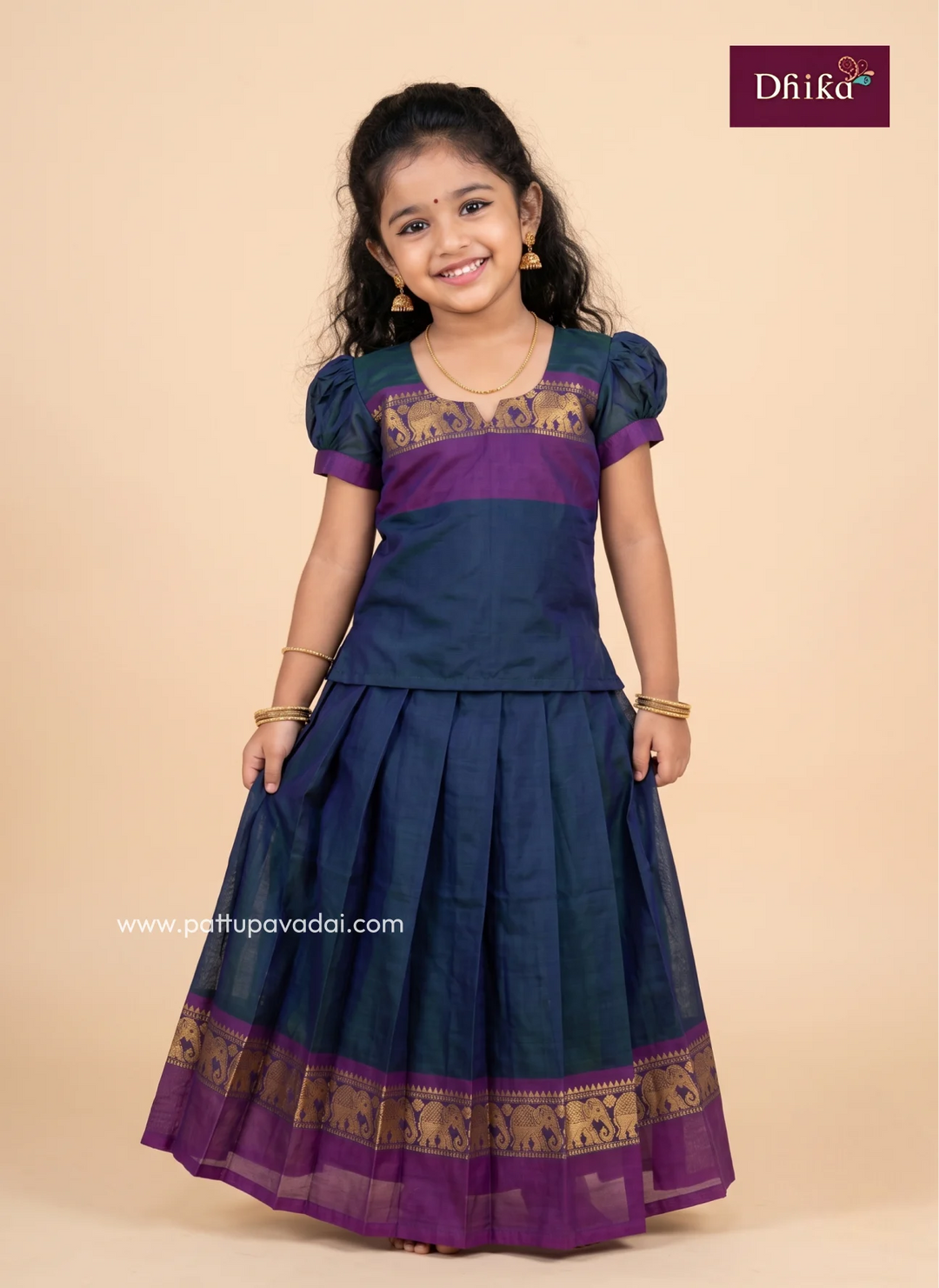 A young girl modeling a traditional Indian dress, featuring a navy blue and purple kurta with gold patterned borders and a matching pleated skirt, set against a solid beige background.
