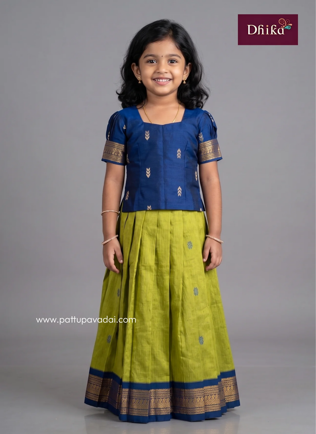 A young girl modeling a traditional Indian outfit consisting of a royal blue short-sleeve top with gold zari embroidery and a matching olive green skirt with a blue and gold border. The image is set against a solid gray studio background with the 'DhiRa' brand logo in the top right corner.