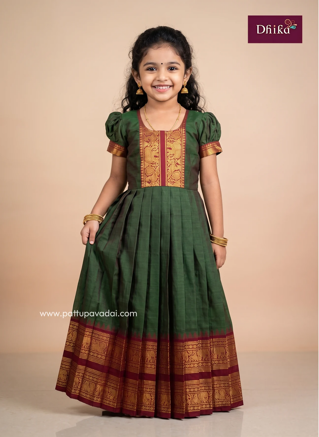 A young girl modeling a dark green traditional Indian dress with gold and maroon zari embroidery, standing against a solid beige studio background. The brand logo 'Dhika' is visible in the top right corner.