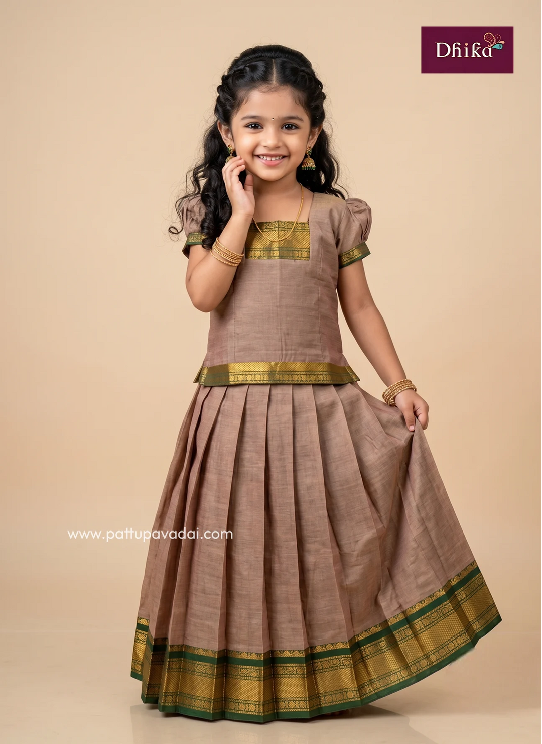 A young girl modeling a traditional Indian dress, featuring a brown pleated skirt and bodice with gold and green borders, set against a solid beige background. The 'Dhika' brand logo is visible in the top right corner.