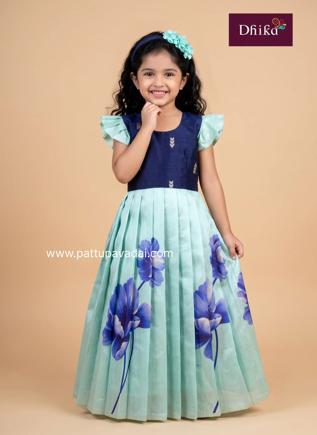 A young girl modeling a traditional Indian dress with a navy blue bodice and a pleated mint green skirt featuring blue floral prints, set against a solid beige background.