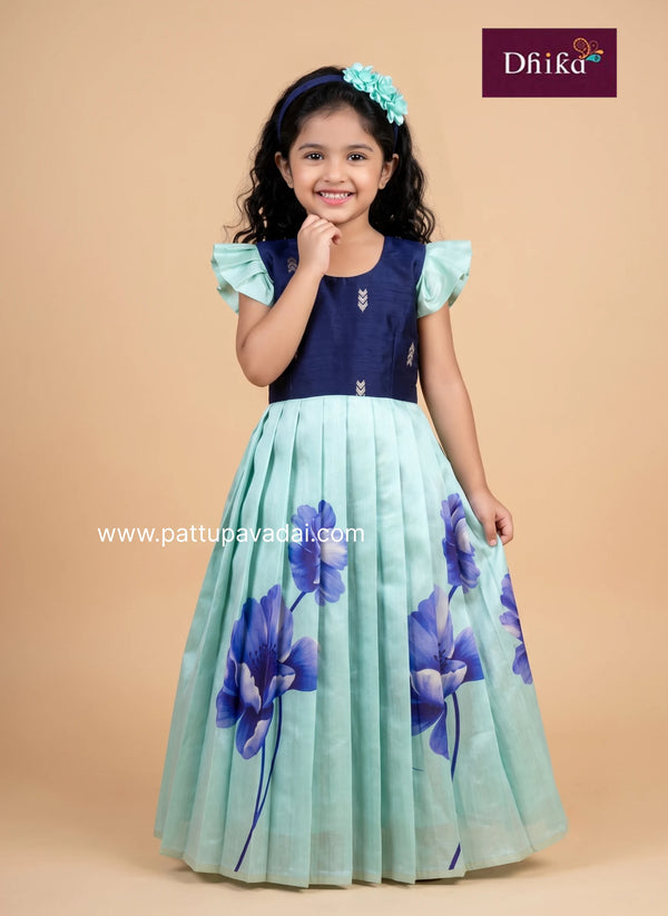A young girl modeling a traditional Indian dress with a navy blue bodice and a pleated mint green skirt featuring blue floral prints, set against a solid beige background.