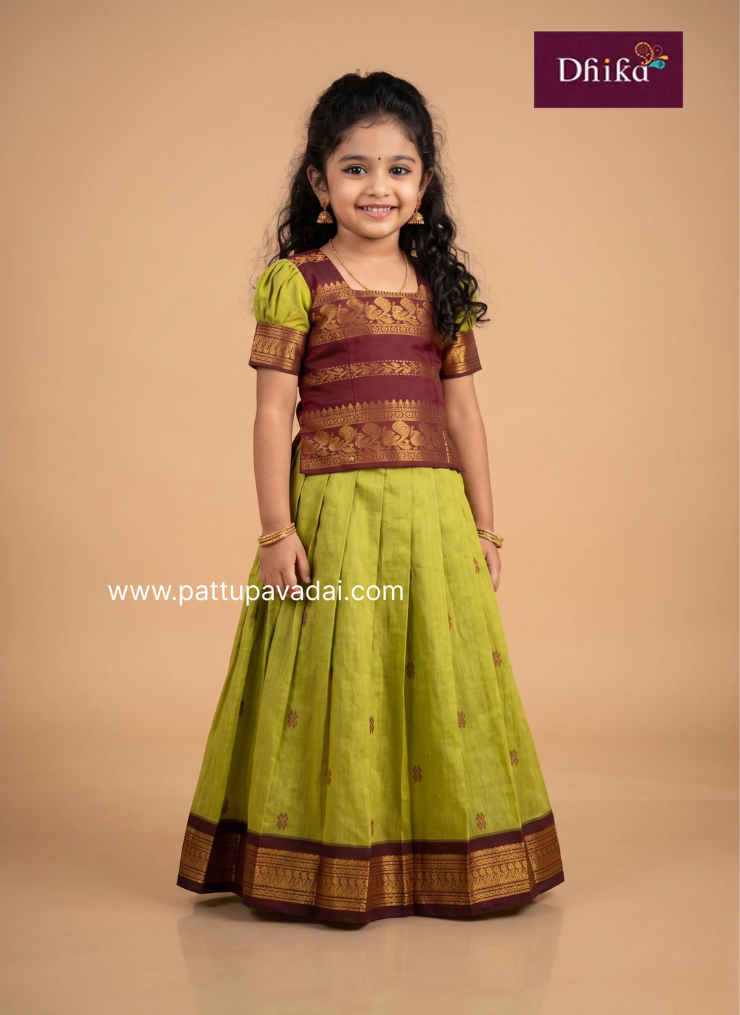 A young girl modeling a traditional Indian ethnic outfit consisting of a lime green skirt with a dark brown and gold zari border and a matching brown and gold embroidered blouse, set against a solid beige studio background. The 'Dhika' brand logo is visible in the top right corner.