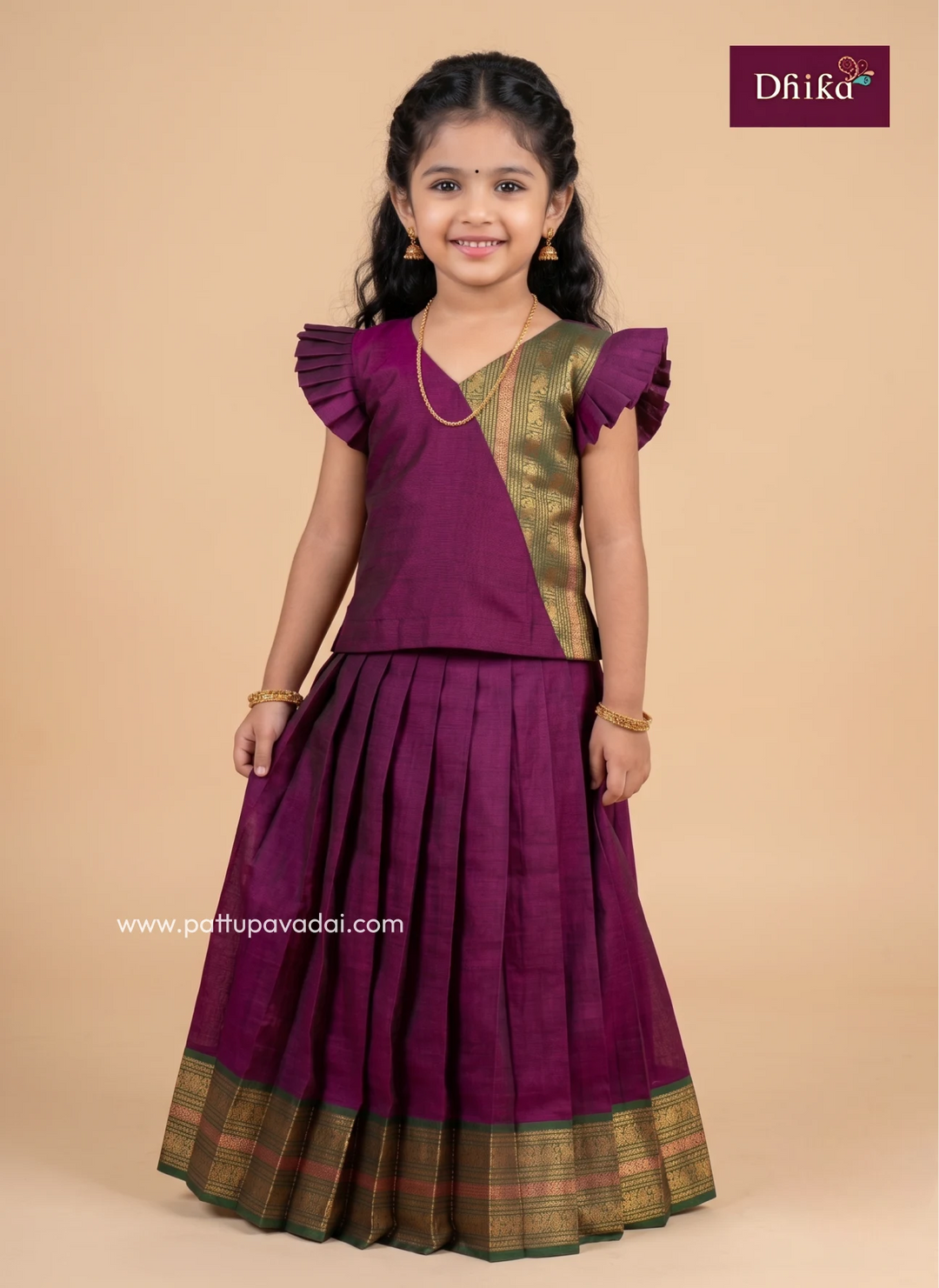 A young girl modeling a traditional Indian outfit consisting of a deep purple skirt with a gold and green border and a matching purple top with gold accents, set against a solid beige background. The 'Dhika' brand logo is visible in the top right corner.