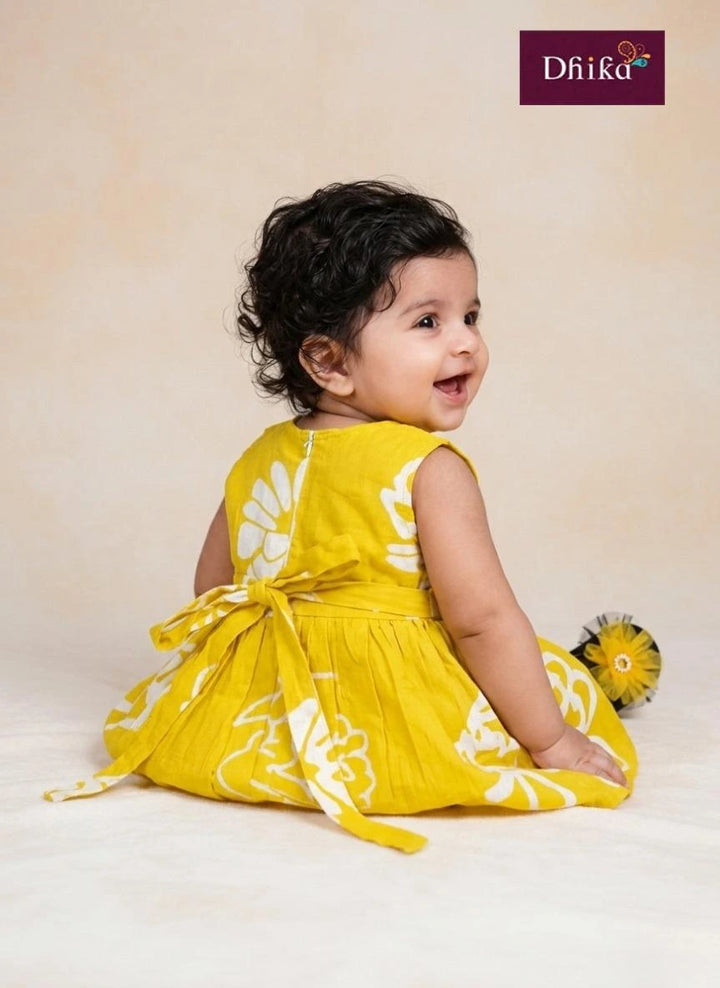 Baby in a yellow dress with white patterns sitting on a white surface, with 'Dhika' branding in the corner.