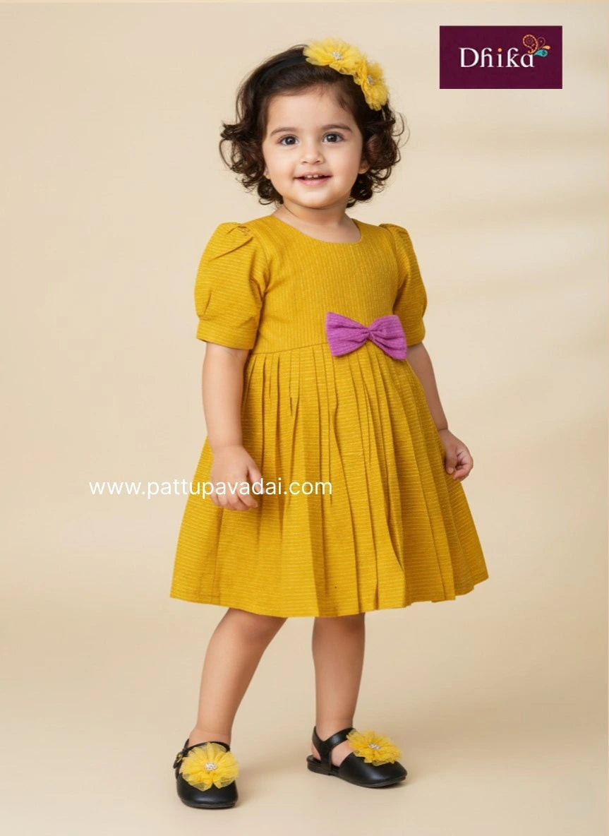 Young girl wearing a yellow dress with a pink bow, standing against a beige background.