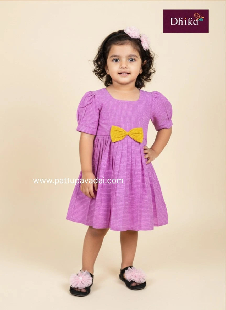 Young girl wearing a purple dress with a yellow bow, standing against a beige background.