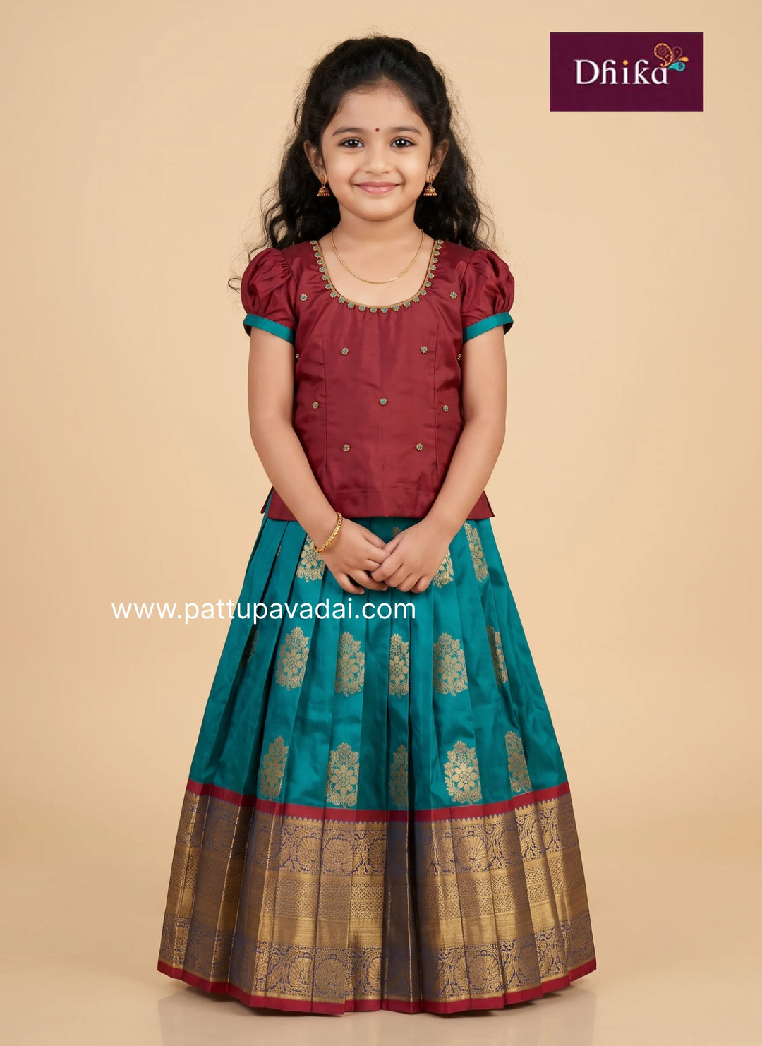 A young girl modeling a traditional Indian ethnic wear set consisting of a maroon embroidered blouse and a teal blue skirt with gold zari patterns and borders, set against a solid beige background. The brand logo 'Dhika' is visible in the top right corner.