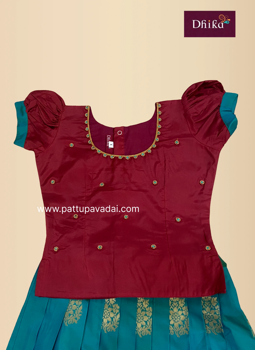 A traditional Indian girl's outfit consisting of a deep maroon satin top with gold embroidery and puff sleeves, paired with a teal blue skirt featuring gold zari patterns. The brand logo 'Dhika' is visible in the top right corner.