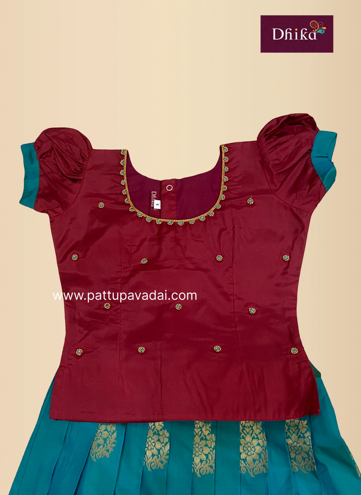 A traditional Indian girl's outfit consisting of a deep maroon satin top with gold embroidery and puff sleeves, paired with a teal blue skirt featuring gold zari patterns. The brand logo 'Dhika' is visible in the top right corner.
